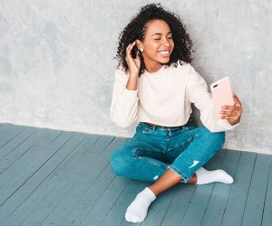 Beautiful black woman with afro curls hairstyle.Smiling model in white trendy sweater. Sexy carefree female sitting near gray wall in studio interior.She taking selfie photos for instagram